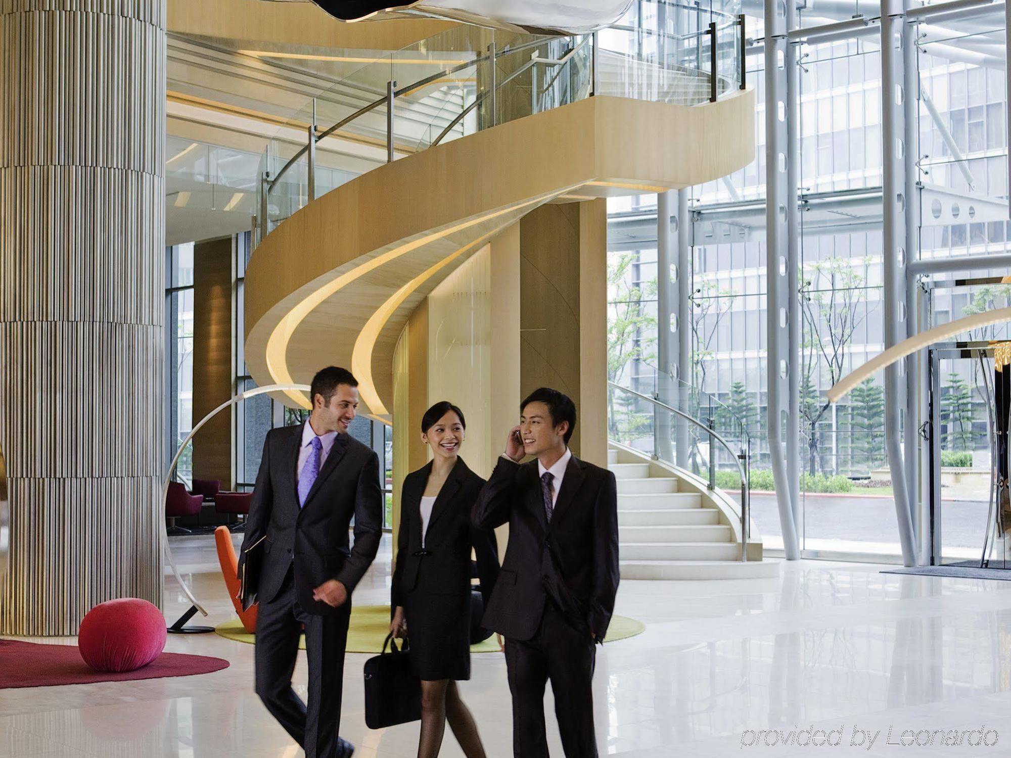 Hyatt Regency Taoyuan International Airport Exterior foto The photo depicts a modern and elegantly designed lobby area with a large, spiraling staircase in the background. Three individuals are walking together, dressed in formal business attire. The setting features large glass windows that allow natural l